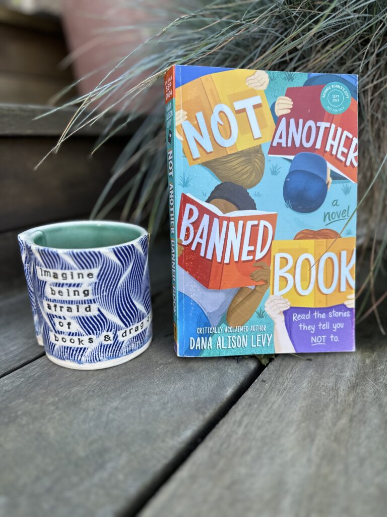 Copy of the book Not Another Banned Book next to a mug that says "Imagine being afraid of books and drag queens"